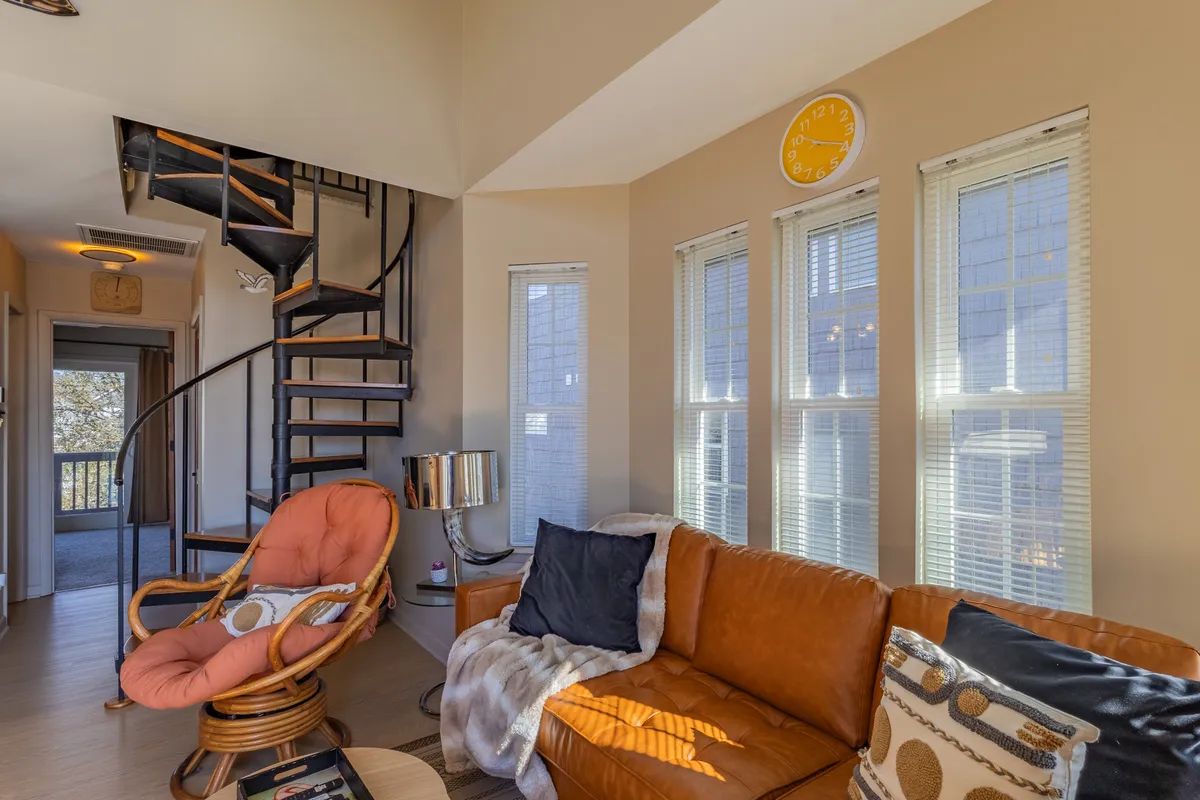 Another inviting view of the living area, spotlighting a spiral staircase, an orange armchair, a leather sofa, and broad windows. The setup feels cozy yet spacious, with seating that encourages togetherness and windows framing the outside world.
