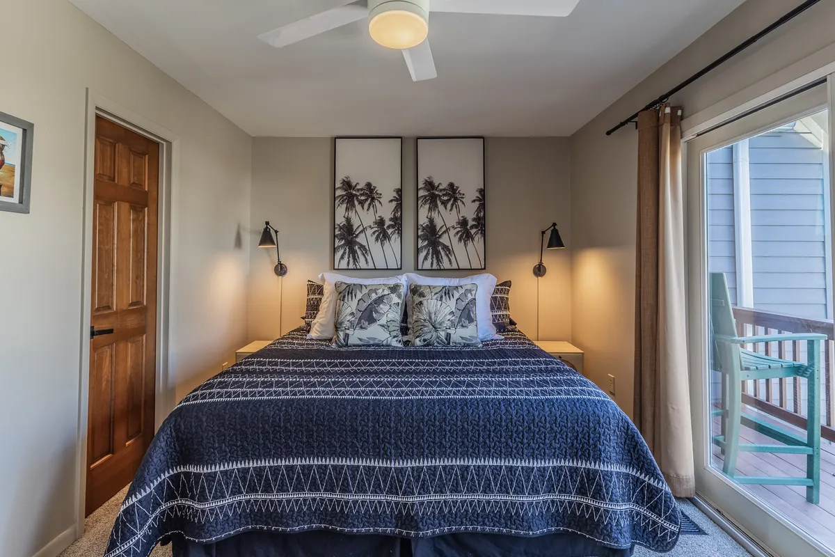 A serene bedroom with a king-sized bed, palm tree artwork above the headboard, matching nightstands, and a sliding glass door that allows sunlight to pour in. The ambiance is cozy and calming, perfect for unwinding at the end of the day.