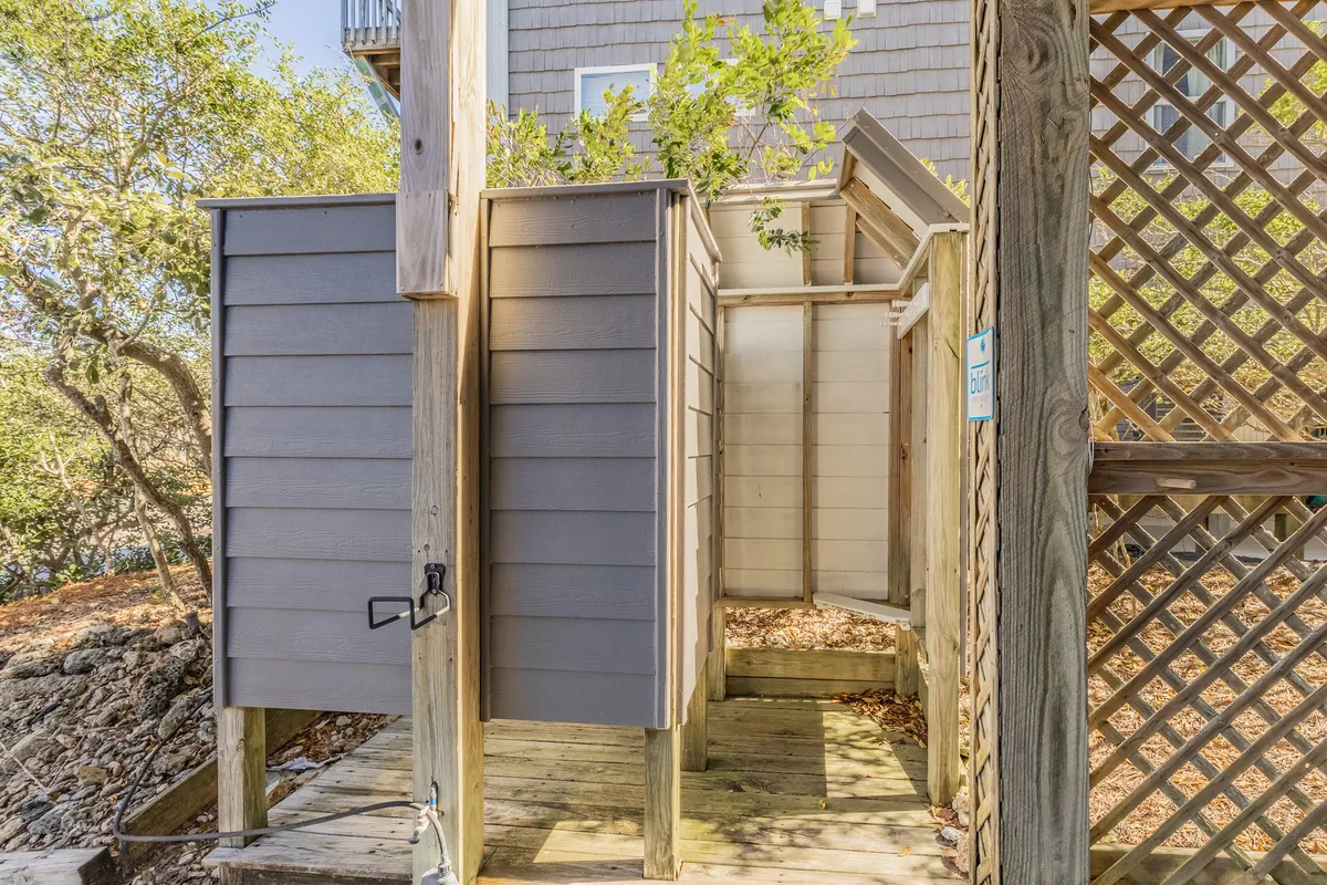 An outdoor shower area with wooden stalls and decking, surrounded by lush greenery for a secluded, nature-inspired feel. The setup promises refreshment and privacy after a day at the beach.