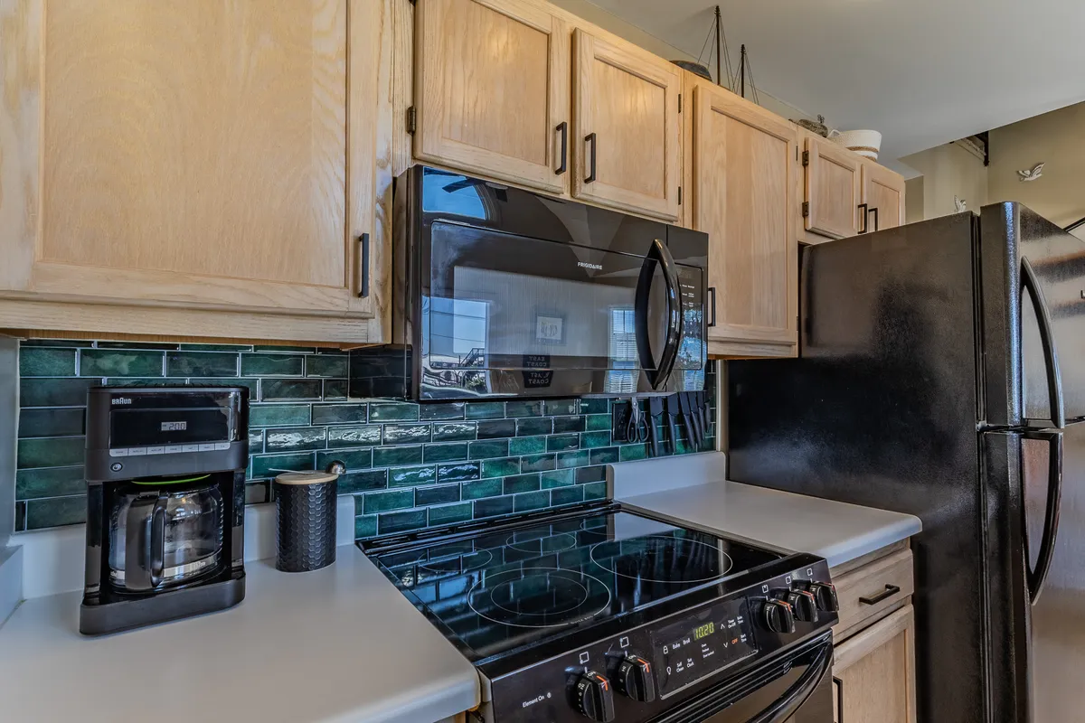This image presents a kitchen scene with black appliances, a green tile backsplash, and warmly toned wooden cabinets. The setup is intimate, cozy, and clearly designed for both cooking and small gatherings, with a layout that encourages conversation and togetherness.