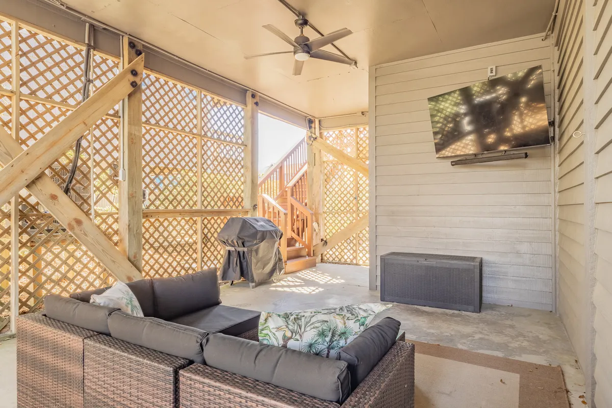 A cozy and spacious outdoor living room featuring a plush sectional sofa, an outdoor TV mounted above, and a grill nearby. The area is shaded and arranged to encourage both relaxation and social gatherings, providing a seamless indoor-outdoor living experience.