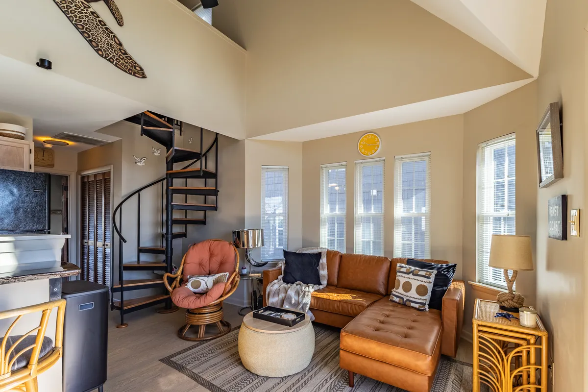 A spacious, sunlit living room featuring a contemporary spiral staircase, plush leather couch, colorful armchair, and expansive windows that fill the area with natural light. The large windows offer stunning views and help blend the indoor space with the scenic outdoors.