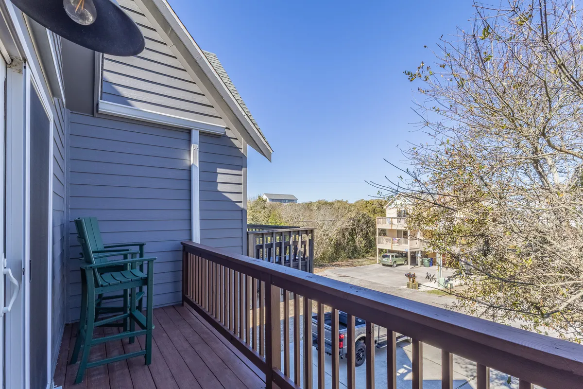 A bright, open balcony features green chairs and a clear blue sky overhead, framed by nearby trees. The space suggests a perfect setting for morning coffee, casual meetings, or simply enjoying the fresh air while working or relaxing.