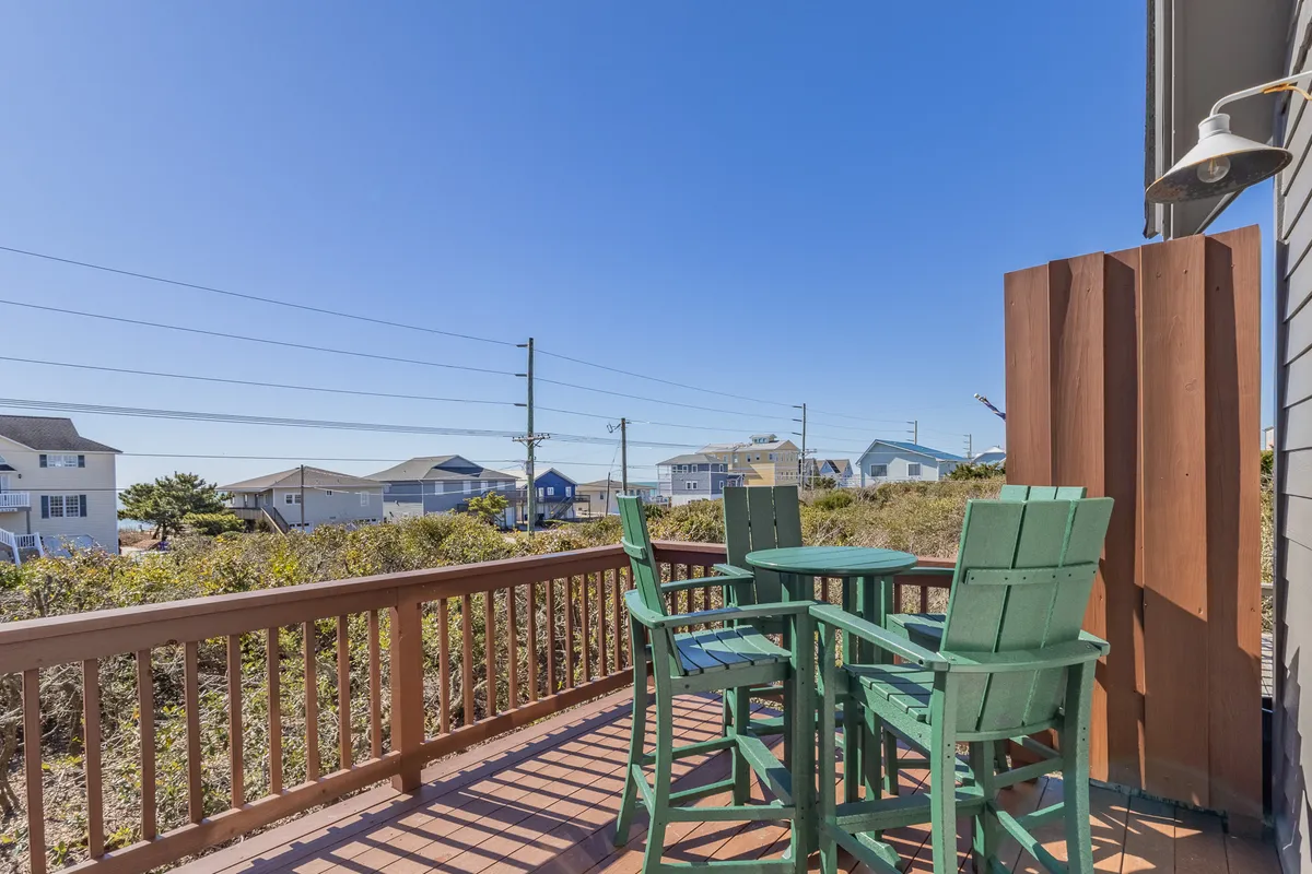 A welcoming outdoor seating area with green chairs and a wooden railing overlooks distant beach houses. The scene suggests a perfect spot for sunrise coffee, evening gatherings, or intimate conversations with friends, encapsulating both privacy and sociability.