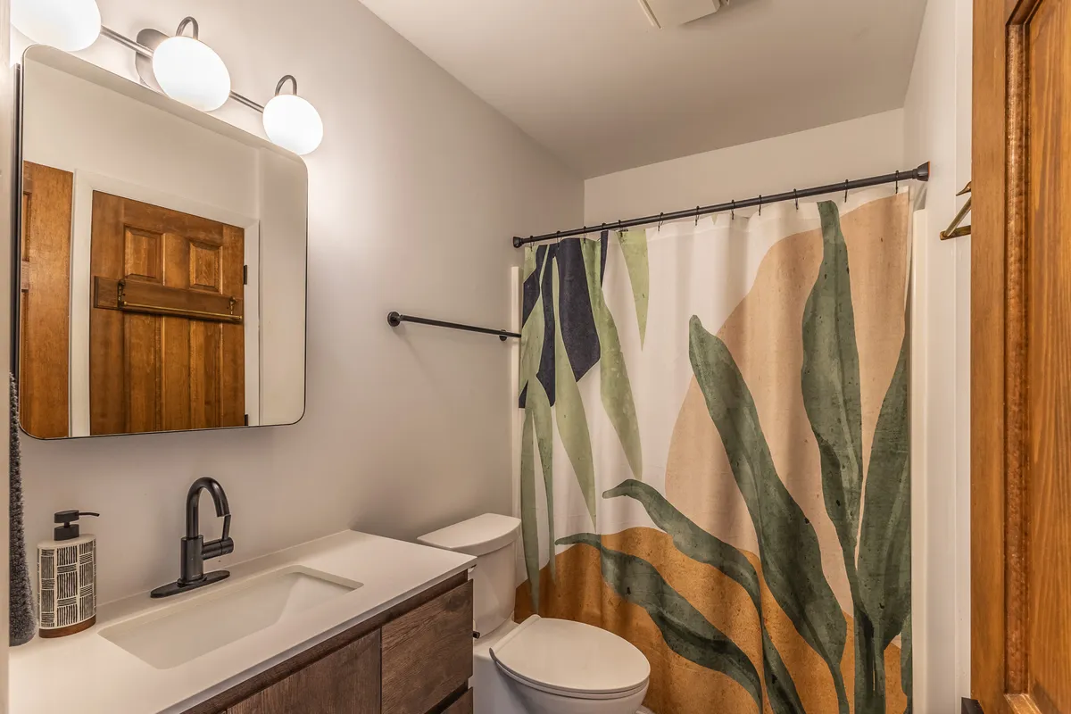 A well-lit bathroom featuring a large mirror, a plant-themed shower curtain, and a wooden door. The space feels fresh, inviting, and ideal for a relaxing bath or refreshing shower at any time of day.