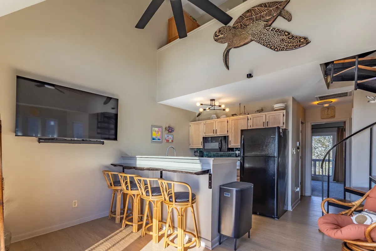 The image showcases a modern kitchen with a spacious island, bar stools for casual seating, a TV for entertainment, wooden turtle wall art, and a striking spiral staircase. The layout is open, with ample room for cooking and socializing, and the decor feels relaxed and welcoming.