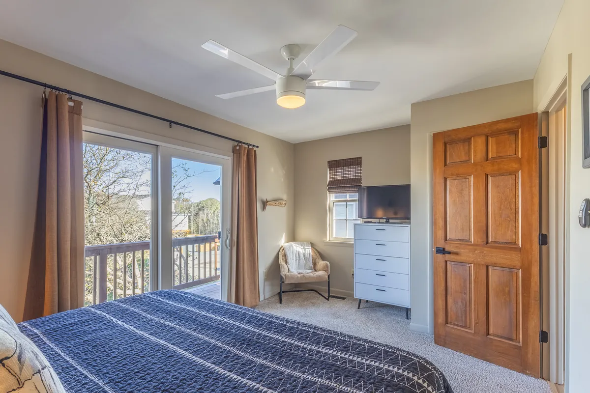 A cozy bedroom featuring a bed with patterned bedding positioned near large windows that offer a beautiful view. The setup includes a dresser with a TV and a comfortable armchair by the window, inviting relaxation and moments of quiet.