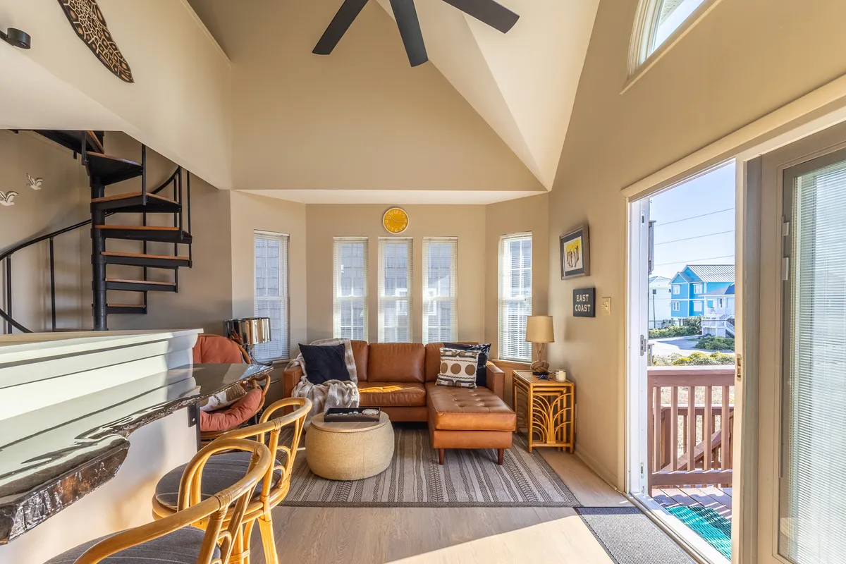 A welcoming interior space with a leather sofa, spiral staircase, large windows, and stylish bar stools. The arrangement encourages both relaxation and conversation, with natural light streaming in and offering glimpses of the outdoors.