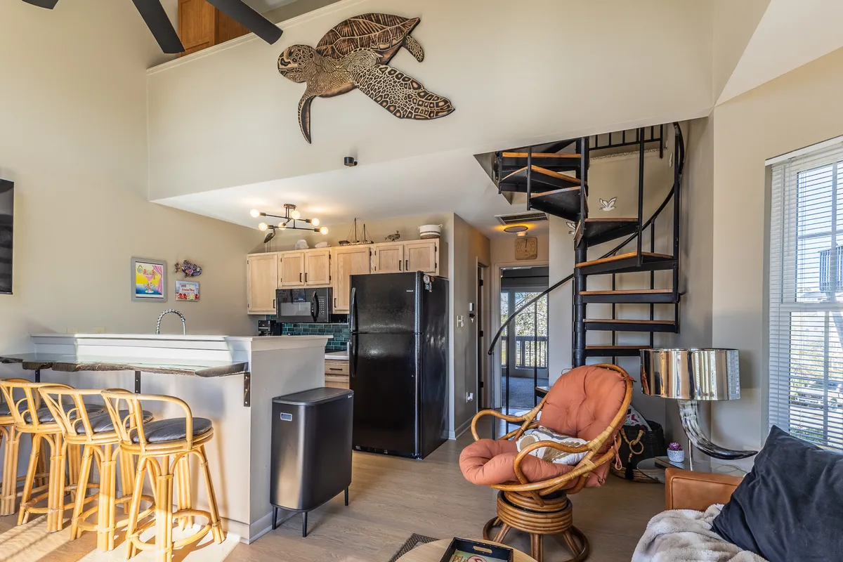 A view of a comfortable dining area featuring a spiral staircase, inviting bar stools, turtle wall art, and a plush chair. Soft natural light filters into the space, and the decor is both artistic and welcoming—ideal for relaxed meals or morning coffee.
