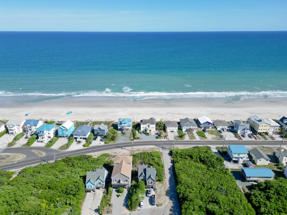 The scene showcases a peaceful stretch of ocean, sandy beach, nearby houses, a road, and coastal vegetation. Families and children can be pictured enjoying beach activities such as building sandcastles, swimming, and relaxing by the surf.